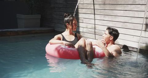 Sunlit Backyard Pool Romance: Asian Woman on Pink Inflatable Ring Chatting with Man