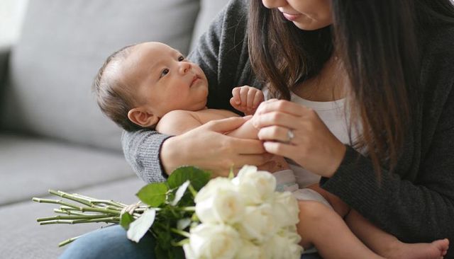 Tender mother cradling newborn on grey sofa with white roses, intimate bonding moment