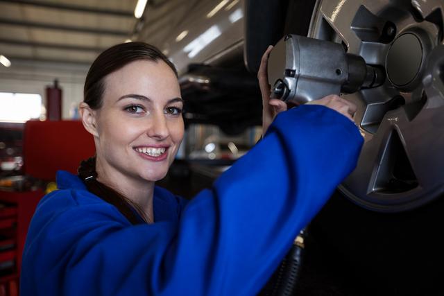 This image shows a female mechanic using a pneumatic wrench to fix a car wheel in an auto repair shop. The mechanic is smiling, conveying professionalism and confidence. This visual can be used for promoting automotive repair services, highlighting gender diversity in the automotive industry, or illustrating professional car maintenance and repair themes.