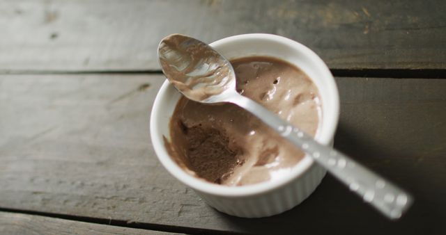 A partially eaten serving of creamy chocolate ice cream is placed in a white ramekin. A metal spoon rests on the edge, indicating the dessert has just been enjoyed. This image shows the contrast between the cool, smooth sweetness of the ice cream and the rustic texture of the wooden table. Perfect for use in recipe blogs, food articles, summer dessert promotions, and advertisements for sweet treats.