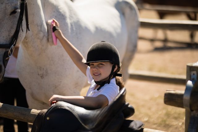 Young girl wearing helmet grooming horse with brush at ranch on sunny day. Ideal for use in materials related to equestrian activities, children's outdoor hobbies, animal care, and rural lifestyle.