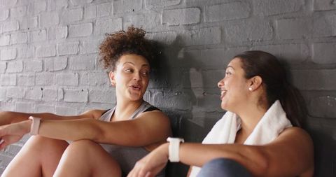 Two diverse women chatting after workout in gym by brick wall with towel, smartwatches