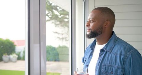 African american man standing by sliding door holding glass of water looking out