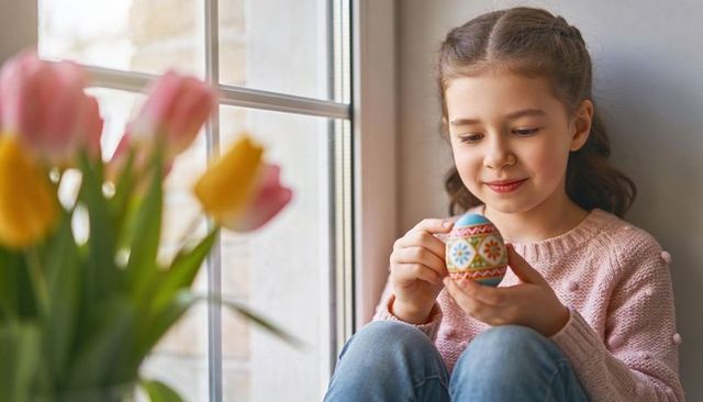 Young girl holding hand-painted easter egg by sunlit window, spring craft moment