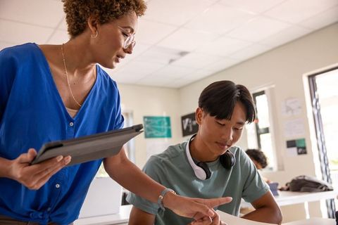 Female teacher guiding teen student with tablet and headphones in sunny modern classroom