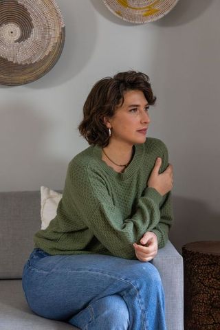 Contemplative woman gazing on gray sofa in green knit sweater and jeans, cozy boho