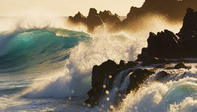 Crashing turquoise wave hitting jagged volcanic rocks in golden backlight