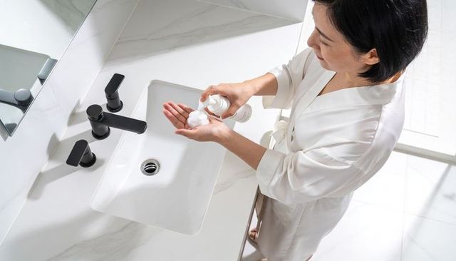 Asian woman dispensing foamy soap at modern marble bathroom sink wearing white robe