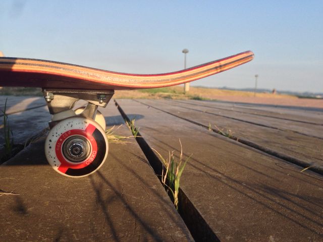 This image captures a close-up view of a skateboard resting on a wooden boardwalk with the sun setting in the background. The focus is on the skateboard's deck and a single wheel, highlighting the texture of the wood and the skateboard design. Ideal for use in articles or promotional materials related to skateboarding, sports, outdoor activities, or lifestyle. Perfect for websites, blogs, magazines, or social media campaigns focusing on youth, adventure, and action sports niches.