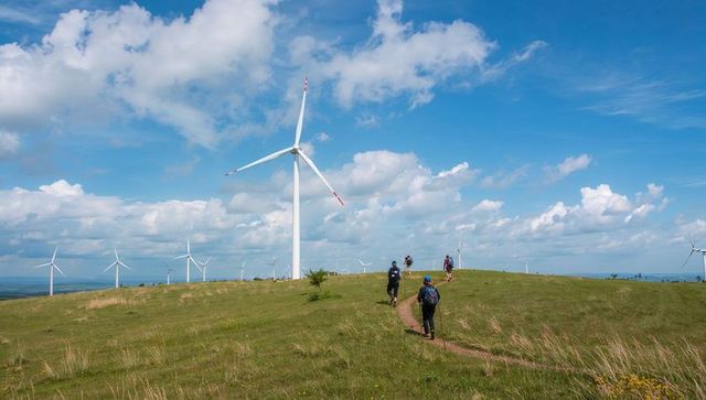 Hikers trekking past towering wind turbine on grassy ridge under blue sky