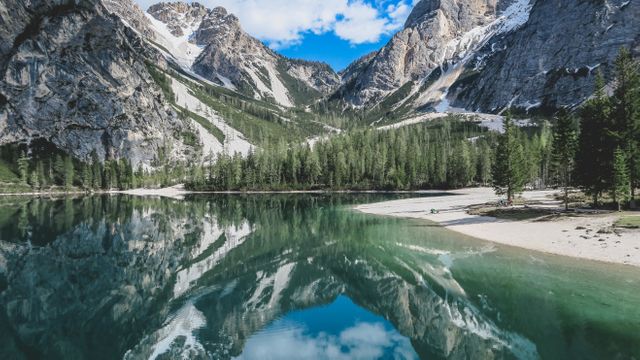 Stunning image of a serene mountain lake with crystal clear water reflecting nearby snow-capped peaks and pine forest. Ideal for use in travel brochures, nature publications, and posters to promote tranquility and outdoor adventure.