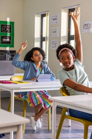 African american preteen girls raising hands in classroom using tablet and notebooks