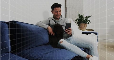 Smiling man relaxing on blue couch using smartphone while petting dog in modern living room
