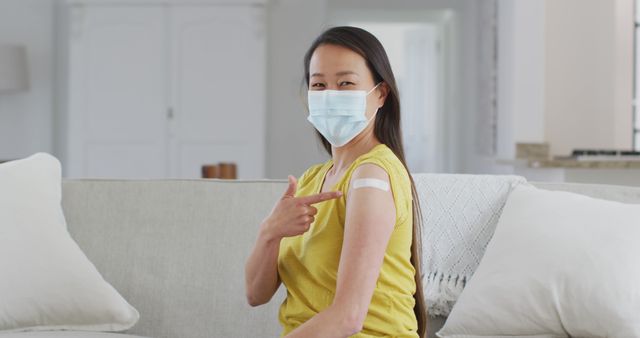 Woman with face mask sitting on sofa showing band-aid after vaccination signifies health and safety during pandemic. Useful for medical campaigns, articles about COVID-19, healthcare promotions.