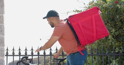 Urban bike courier leaning on bicycle carrying large red delivery backpack on promenade