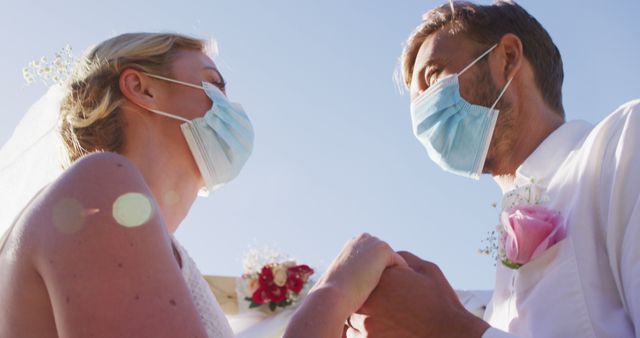 Bride and groom wearing surgical masks holding hands and staring into each other’s eyes during an outdoor wedding ceremony. The image captures a unique combination of love and pandemic precautions. Ideal for articles or materials discussing pandemic weddings, love during COVID-19, or creative ways couples celebrate important milestones safely.