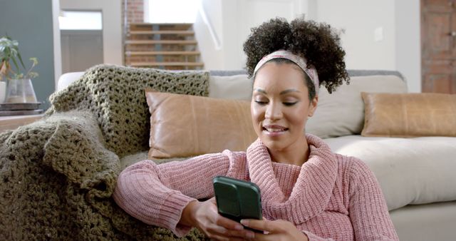 A photo of a smiling woman with curly hair relaxing on a sofa at home and scrolling on her smartphone. She wears a cozy pink knit sweater and a headband. This image can be used in contexts related to home lifestyle, technology use, relaxation, and personal leisure. Ideal for blogs, social media posts, and advertisements related to comfortable living, tech gadgets, or well-being.