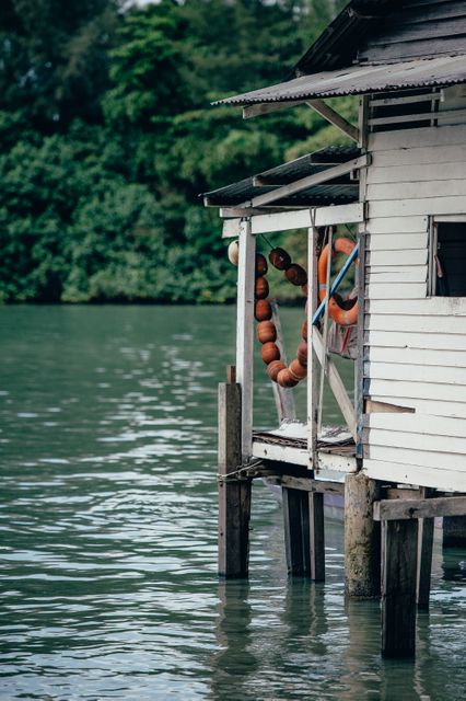 This image shows a wooden house on stilts standing over a tranquil river and surrounded by lush greenery. Life preservers hang along the side of the house, adding a touch of safety and traditional charm. It can be used for marketing travel destinations, promoting rural tourism, illustrating traditional architecture, or evoking a peaceful, back-to-nature lifestyle.