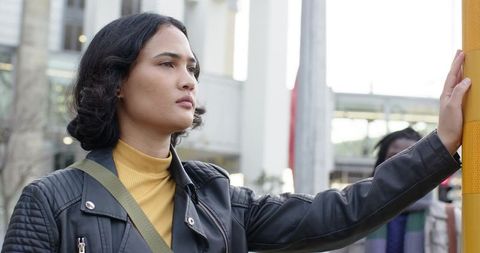 Young commuter standing at bus stop touching yellow pole, looking into urban distance