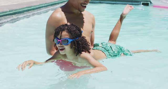 Father giving son swimming lessons in a pool during summer. Son is wearing goggles and green swim trunks, smiling with joy. Ideal for use in content related to family activities, summer fun, swimming tutorials, or promoting swimming safety.