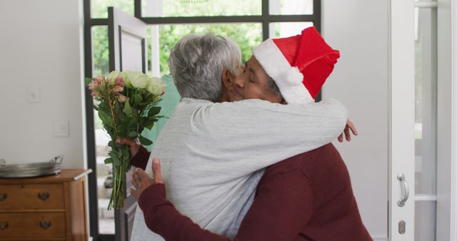 Two elderly women sharing a heartfelt hug, one wearing a Santa hat and holding a bouquet of flowers, conveying holiday spirit and celebration. Scene suggests warmth, friendship, and affectionate reunions. Ideal for themes related to holidays, senior relationships, family gatherings, and festive greeting cards.