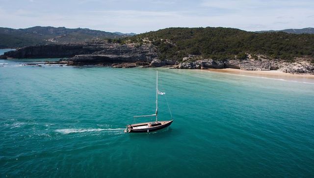 Sailboat sailing turquoise cove leaving wake near sandy beach and rugged limestone cliffs