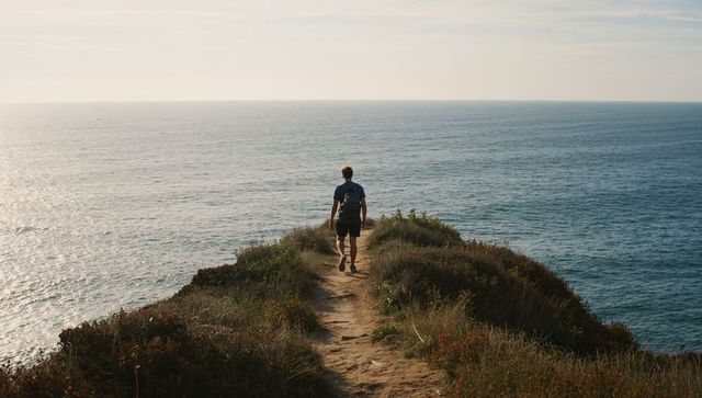 Lone hiker walking coastal bluff trail toward endless ocean horizon at warm golden light