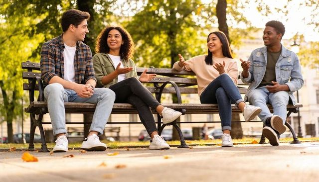 Four friends chatting on sunlit park bench during autumn in urban park, casual sneakers