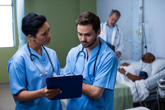 Nurses in blue scrubs discussing patient care while holding a clipboard in a hospital ward. A doctor is attending to a patient in the background. This image can be used for healthcare, medical teamwork, hospital environment, and patient care themes.