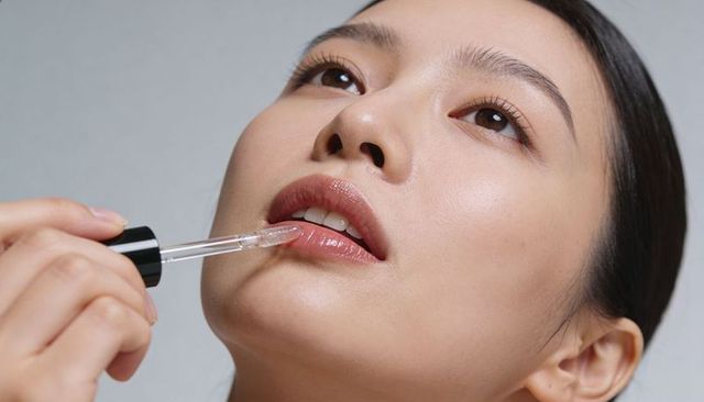 Close-up asian woman applying clear lip gloss with dropper showing dewy natural makeup