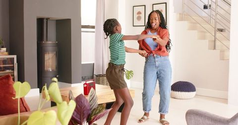 Joyful African mother and teenage daughter dancing and laughing while holding hands at home