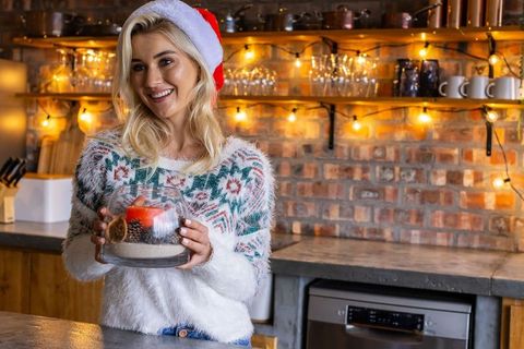 Blonde woman wearing santa hat holding festive centerpiece with candle in rustic kitchen