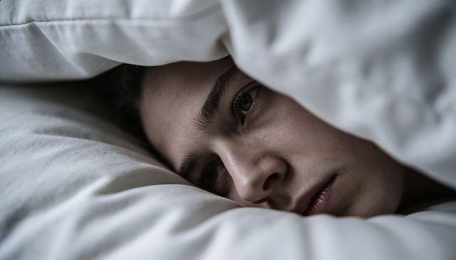 Young man resting under white duvet closeup showing pensive expression and intimate calm
