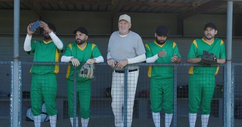 Senior coach leaning on dugout railing with baseball players in green uniforms