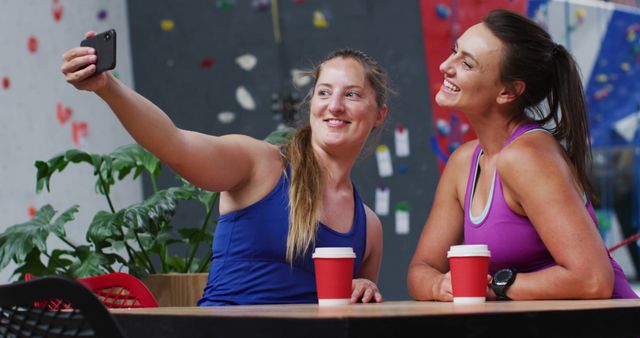 Two women sitting in gym attire taking selfie while enjoying coffee after workout session. Represents friendship, healthy lifestyle, and joy of spending time together. Ideal for use in social lifestyle blogs, fitness commercials, promotional material for gyms, and social media content.