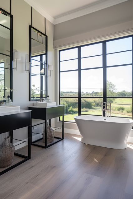 This image portrays a modern bathroom featuring a sleek freestanding tub positioned in front of expansive windows that offer a stunning view of nature. With double sinks on black vanities, ample natural light floods the room, enhancing the minimalist and luxurious ambiance. Ideal for use in home decor websites, interior design blogs, and modern architecture platforms.