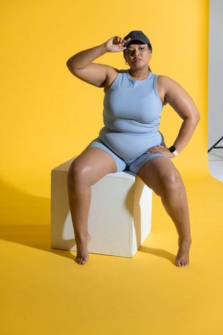 Confident curvy woman wearing blue athleisure sitting on cube with yellow background