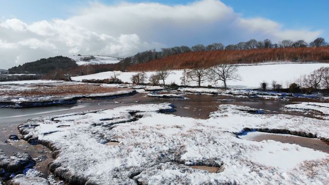 This video of an aerial view shows a serene, snow-covered landscape with a winding river running through it. Trees stand bare against a clear winter sky, with gentle hills in the background. Use this for nature travel blogs, winter holiday promotions, seasonal greetings, or environmental presentations.