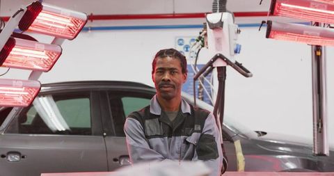 Experienced auto body technician inspecting gray car under red infrared curing lamps
