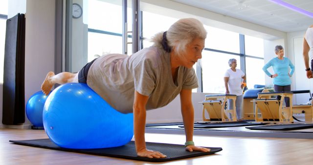 Senior woman engaging in a Pilates workout using a blue exercise ball in a modern fitness studio. Ideal for promoting senior fitness, healthy aging, and rehabilitation exercises. Useful for health, wellness, and fitness industry advertising.