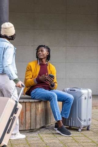 Young travelers conversing at transit terminal sitting with luggage and smartphone