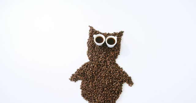 Coffee beans are arranged in the shape of an owl with two cups for eyes, against a white background, with copy space. It's a creative presentation that combines the love for coffee with artistic expression.