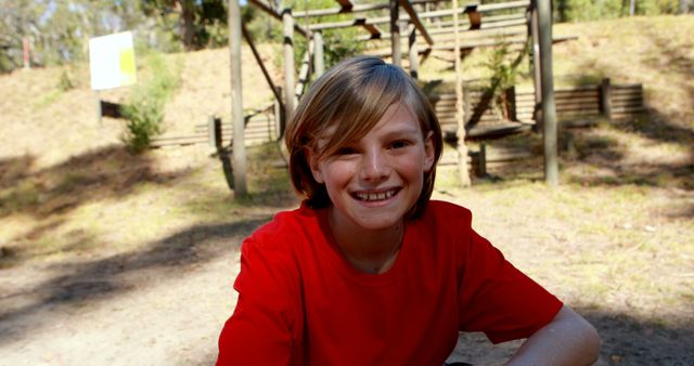 Young boy wearing a red shirt sitting outside at an adventure playground, smiling brightly. Ideal for themes related to childhood, outdoor activities, happiness, play, and family fun in nature settings.