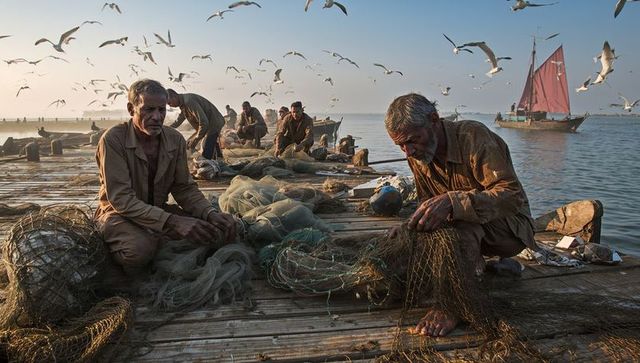 Fishermen mending nets on rustic jetty at sunrise with red-sailed boat and seagulls