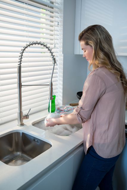 Woman washing bowl under sink in modern kitchen. Ideal for illustrating domestic chores, housework, and daily routines. Suitable for articles on home cleaning tips, kitchen hygiene, and lifestyle blogs.