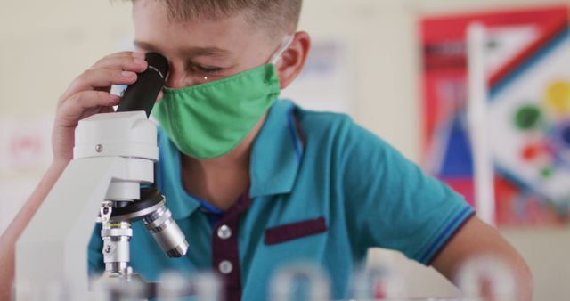 Young boy wearing a green face mask is observing through a microscope in a science classroom. This image illustrates concepts of education, safety, and learning during the pandemic. Ideal for content about scientific education, primary school classroom activities, health and safety measures during Covid-19, and children's engagement in science.