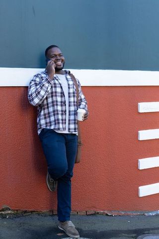 African american man leaning on two-tone wall, talking on phone, holding coffee cup