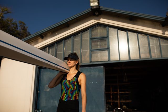 Low angle front view of a Caucasian female rower carrying a boat on her shoulder out of a boathouse with blue sky on a sunny day.