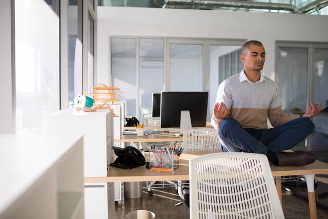 Male executive practicing yoga on desk in a modern office. Ideal for illustrating concepts of workplace wellness, stress relief, mindfulness, and work-life balance. Suitable for use in corporate wellness programs, mental health awareness campaigns, and business lifestyle articles.