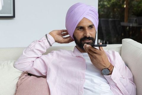 Indian man using smartphone voice function relaxing on sofa